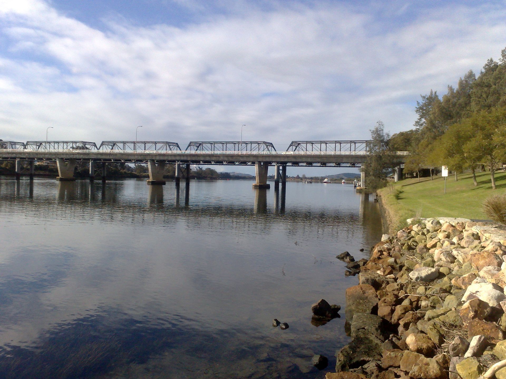 Bridge over water with a cloudy sky. Trees and a grassy bank on the right. Rocky shoreline in the foreground.