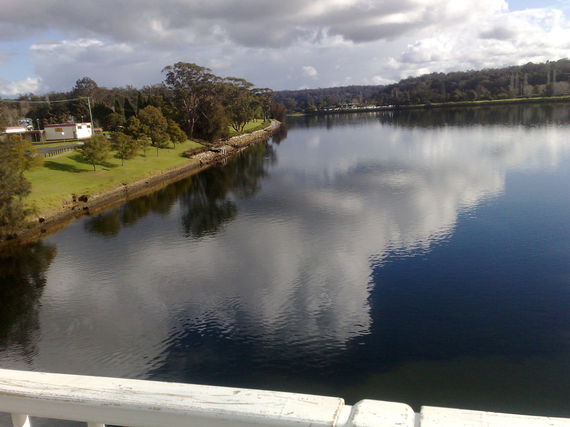 Calm river reflecting cloudy sky and shoreline with trees and buildings.