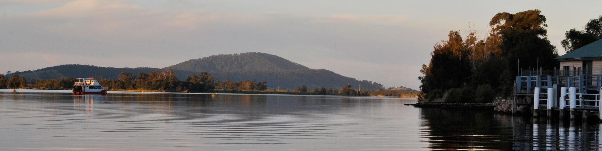 Panoramic view of a calm river with a boat, distant mountains, and a building with a dock.
