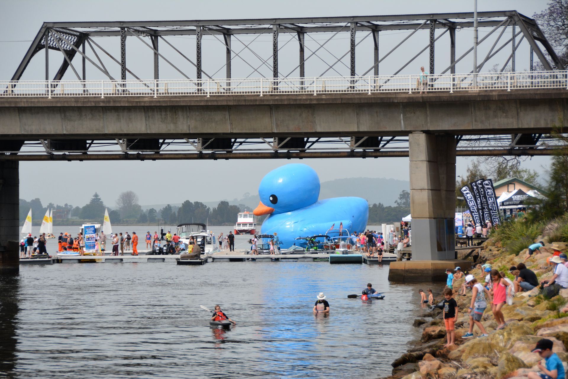 Large blue inflatable duck floats in water under a bridge, people nearby.