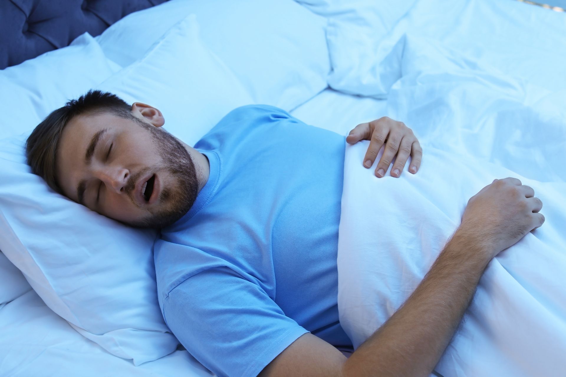 Man sleeping in bed under a white blanket, wearing a blue shirt.
