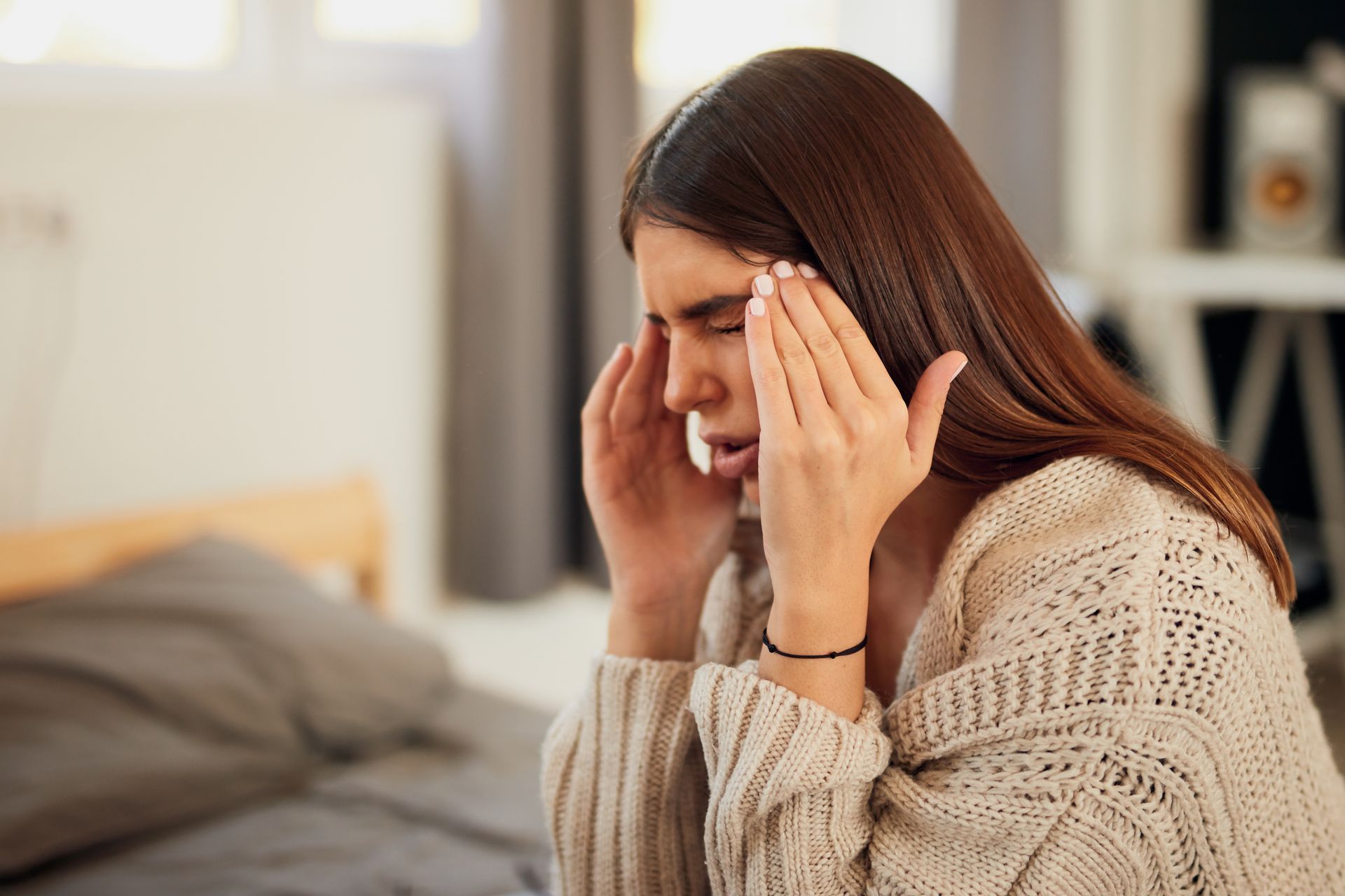 Woman sitting on a bed, holding her temples with a pained expression in a bedroom