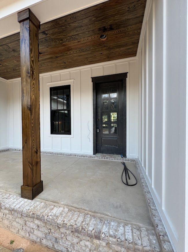 Covered porch with white paneling, a dark door, window, and wood column. Concrete floor, brick base, and stained wood ceiling.
