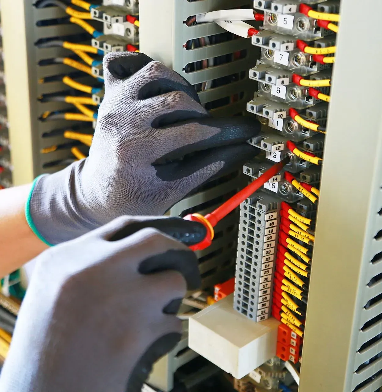 Gloved hands using a screwdriver to wire an electrical panel with red and yellow cables