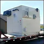 White modular building section on a flatbed trailer, with windows and mounted equipment.