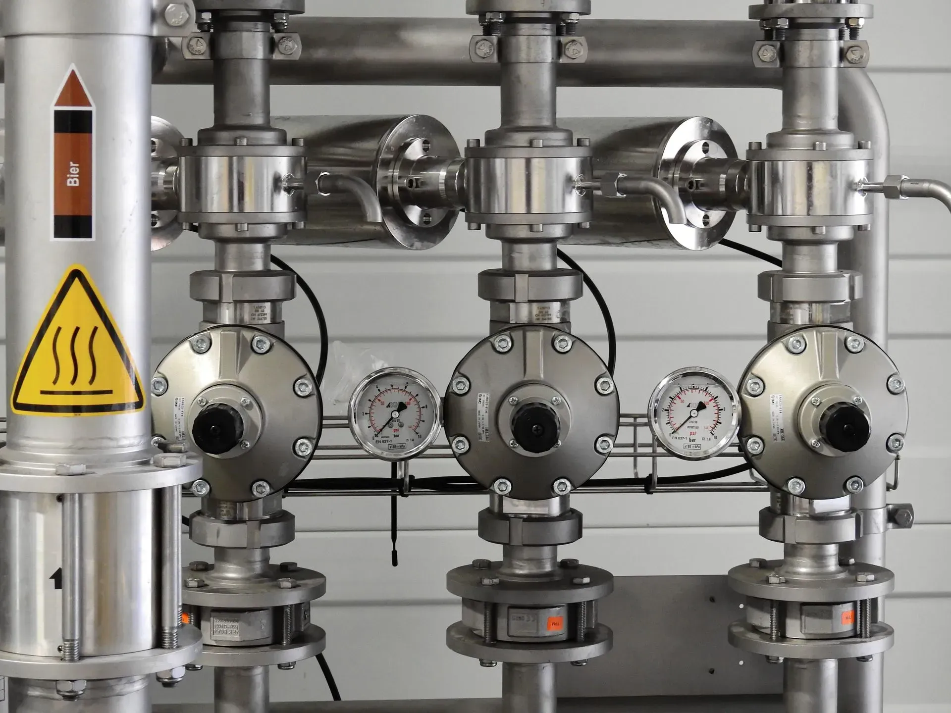 Industrial pipes and gauges in a metal plant, with a yellow heat warning sign on the left.