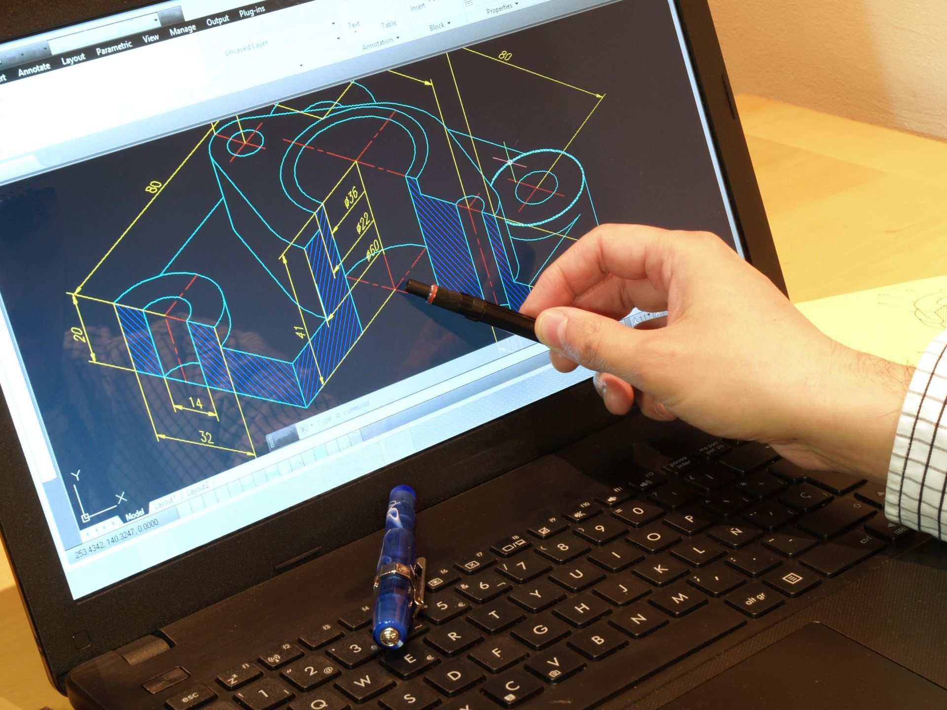 Hand pointing at a CAD design on a laptop screen with a stylus on the keyboard