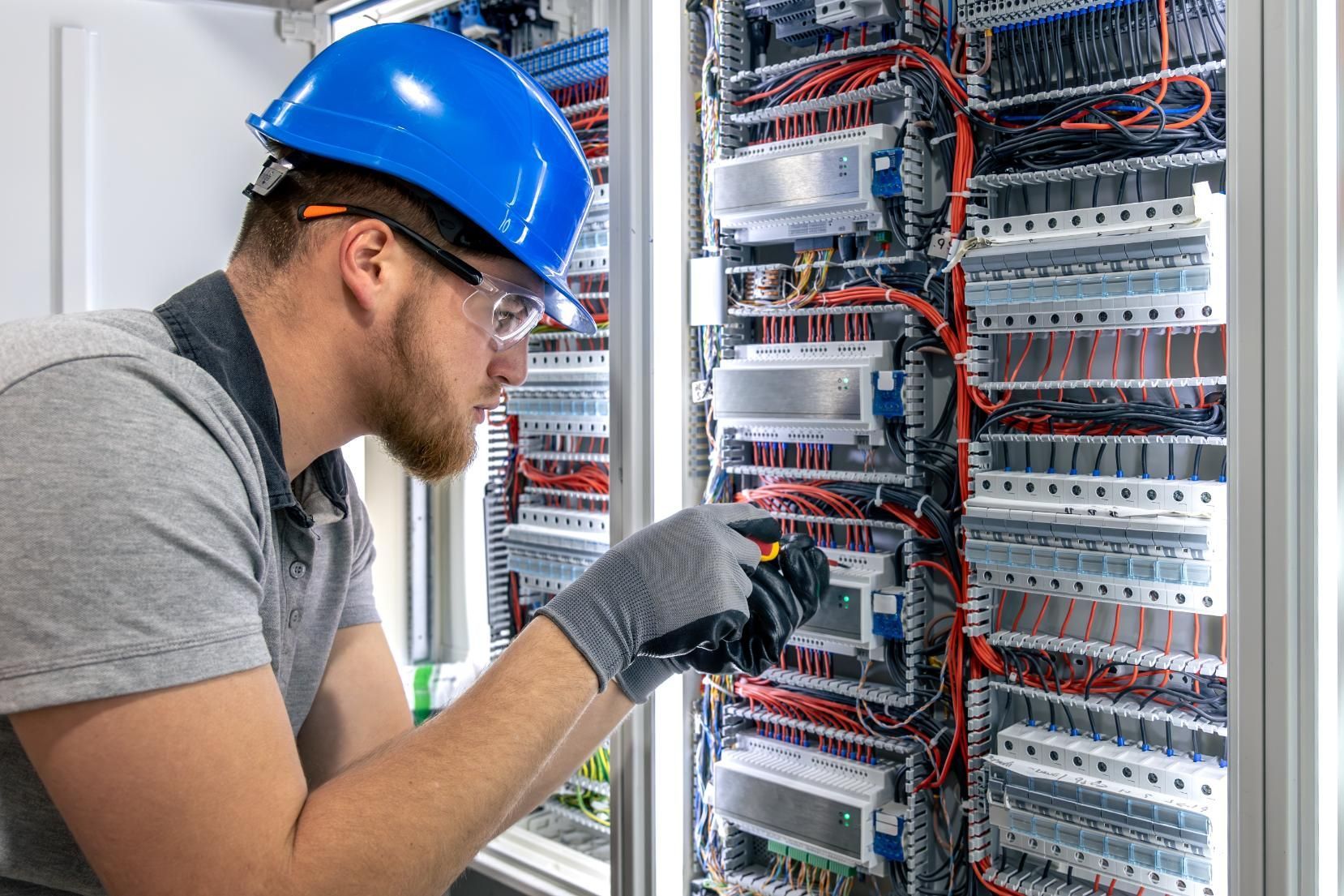 Technician in blue hard hat inspecting wiring inside an open electrical control panel.