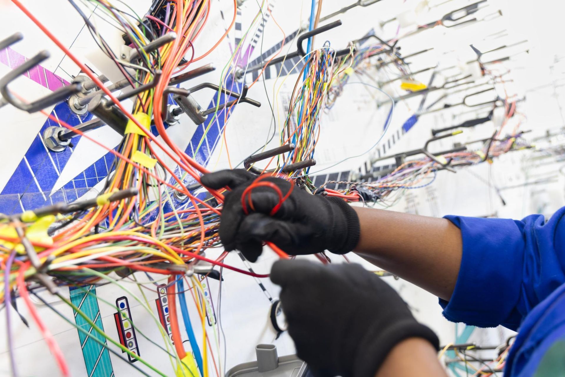 Gloved hands working on a dense tangle of colorful electrical wires on a wall panel
