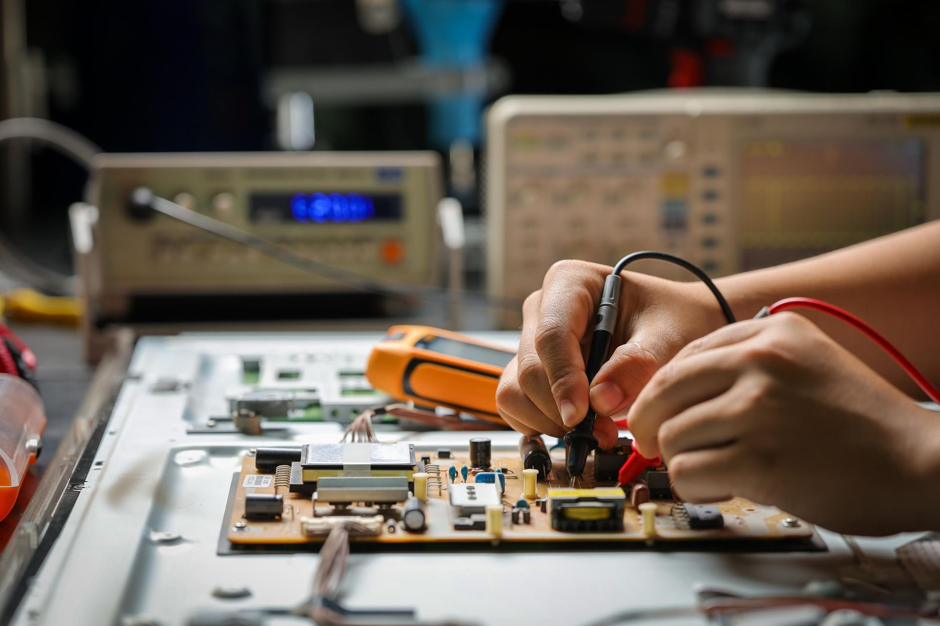 Hands using a multimeter to test a circuit board on a workbench in an electronics repair lab