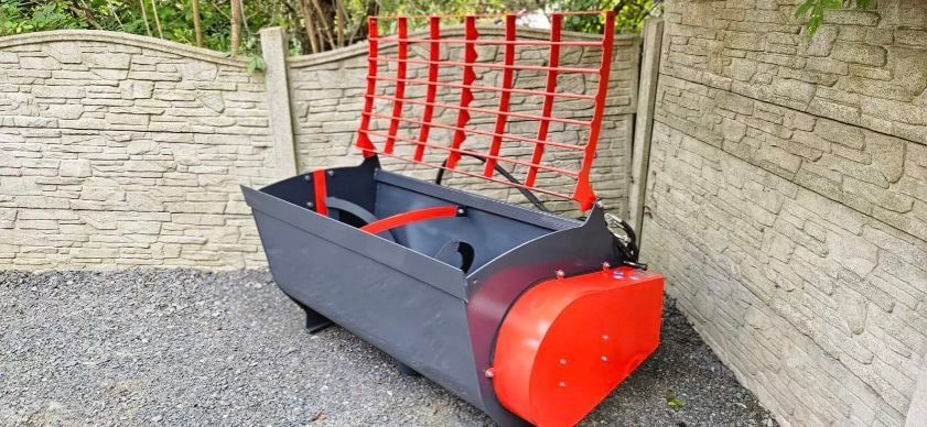 Grauer und roter Futtermischwagen. Er hat rote Zinken und ein rotes Getriebe. Er steht draußen an einer Steinmauer.