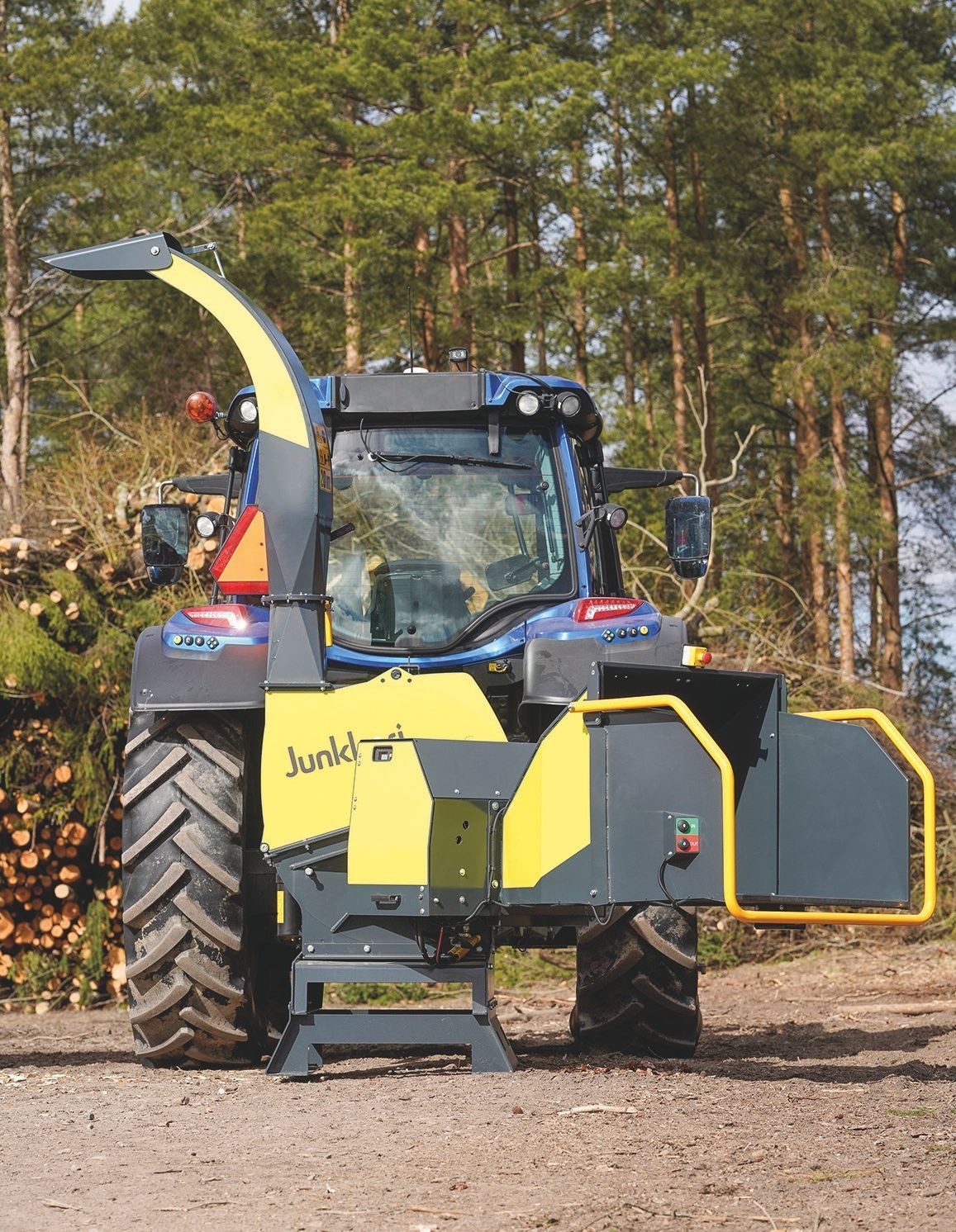 Ein gelb - schwarzer Traktor steht auf einem Feldweg vor einem Stapel Baumstämme.