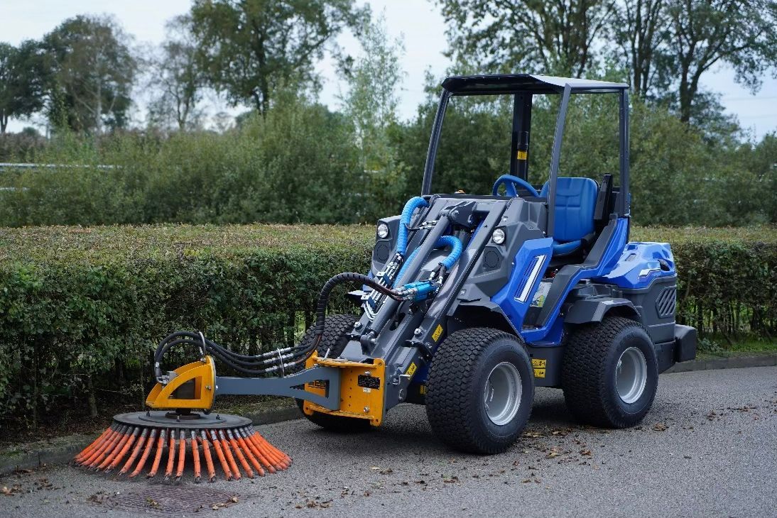 Eine blau-graue Kompaktkehrmaschine kehrt auf einer gepflasterten Fläche Schutt zusammen.