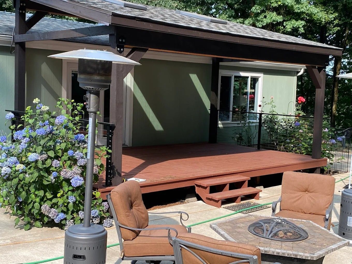 A backyard patio featuring a wooden deck with a covered roof, a fire pit, outdoor furniture, and a large hydrangea bush.