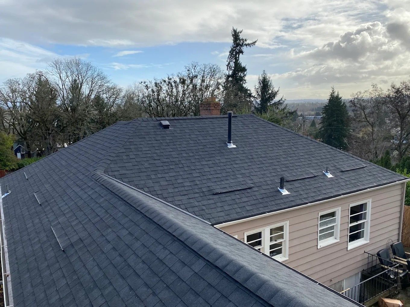 A high-angle view of a newly installed dark grey asphalt shingle roof on a house with light tan siding.