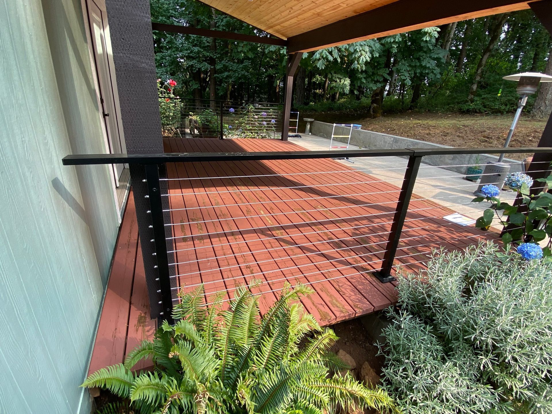 Red wooden deck with a black cable railing overlooking a yard with trees and bushes under a patio cover.
