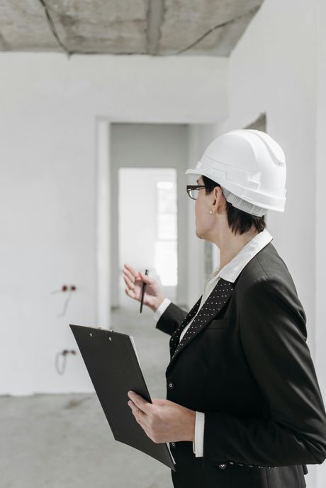 A professional in a hard hat and business suit holds a clipboard and gestures toward a room under construction.