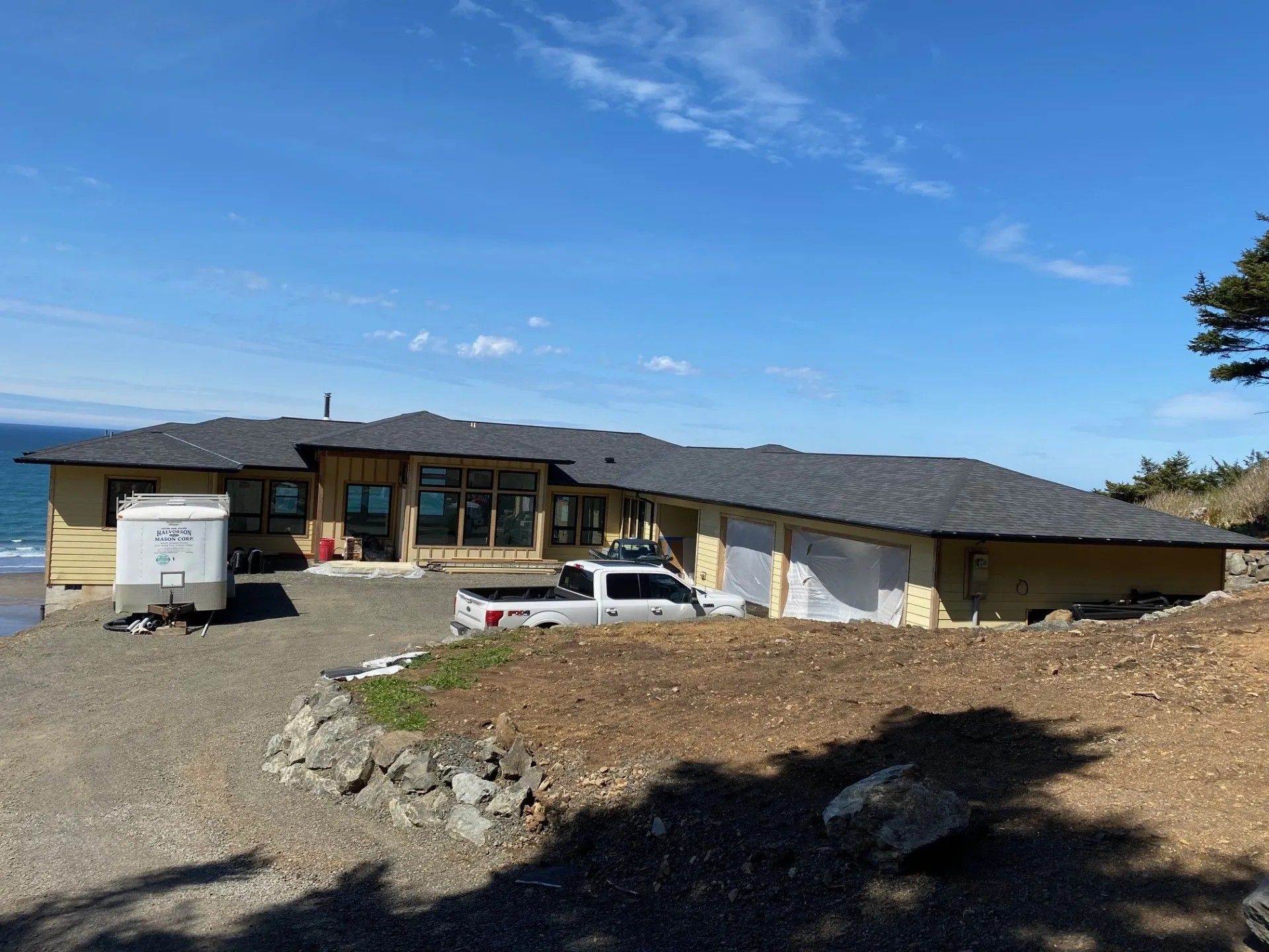 A single-story house with tan walls under construction sits on a gravel lot near the ocean under a clear blue sky.