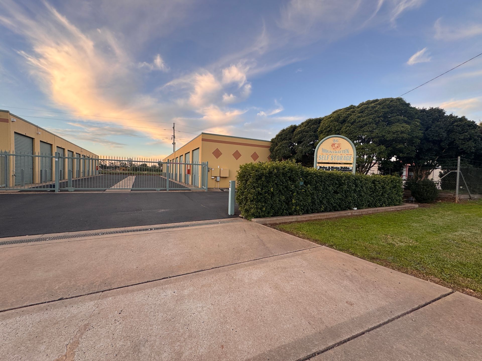Industrial building entrance with gated driveway, hedges, and a sunset sky