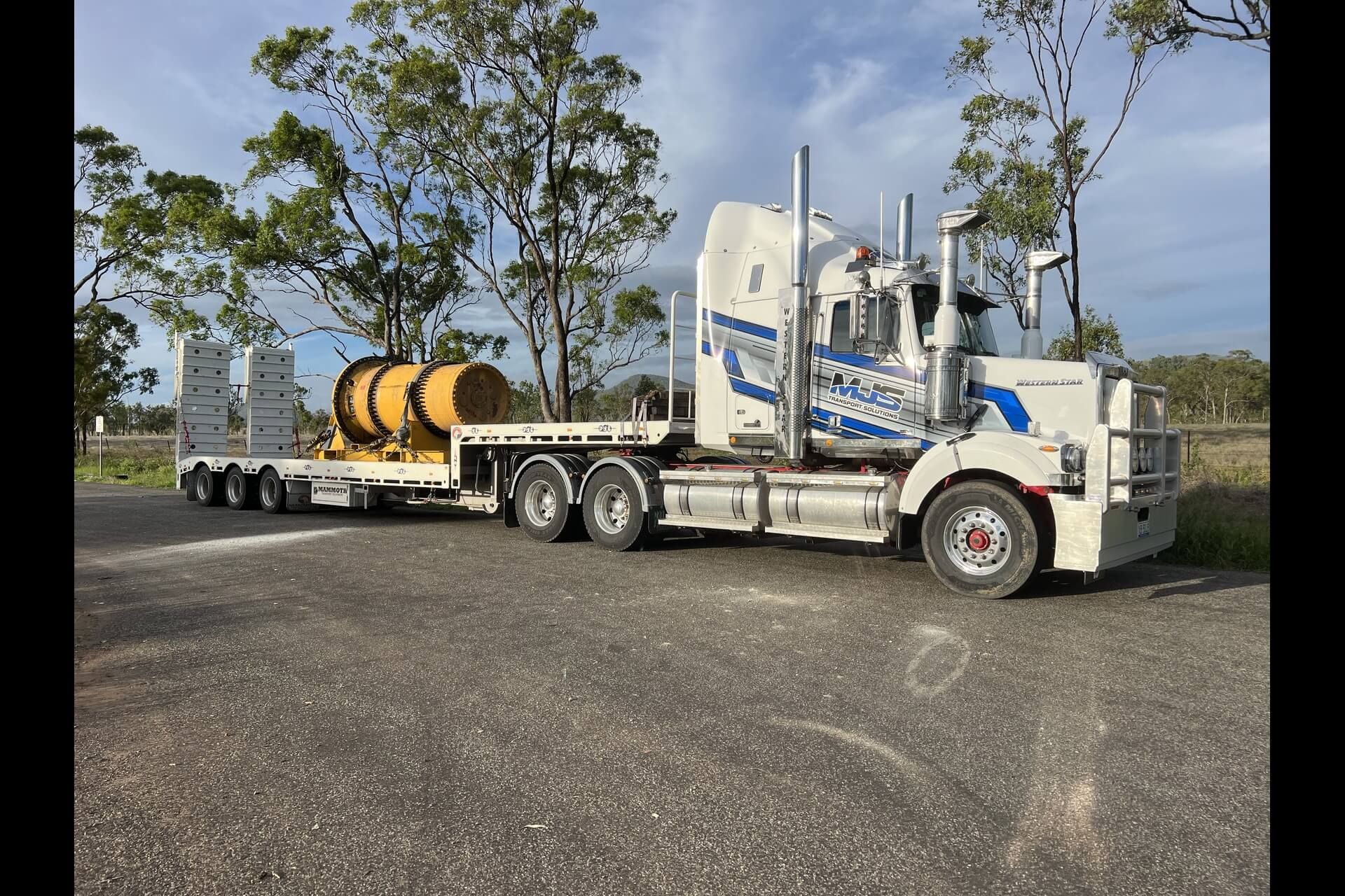 A white semi-truck with blue accents parked on an asphalt surface, pulling a flatbed trailer carrying a large yellow tank.