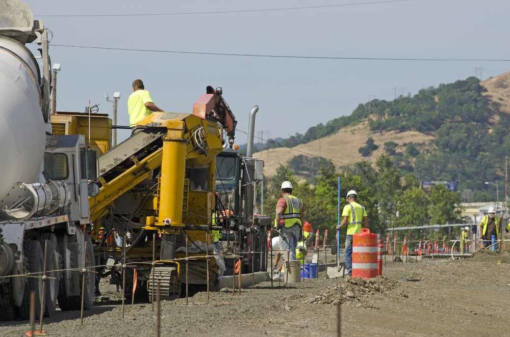 Construction crew working with heavy machinery on a road site with orange traffic barrels and hills in the background