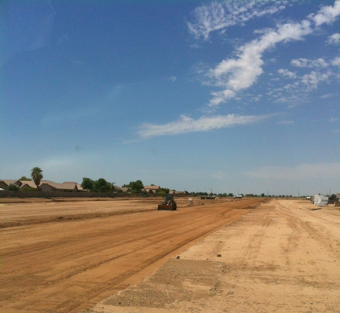 Wide dirt road under a blue sky, with a construction vehicle grading the ground in a rural area.