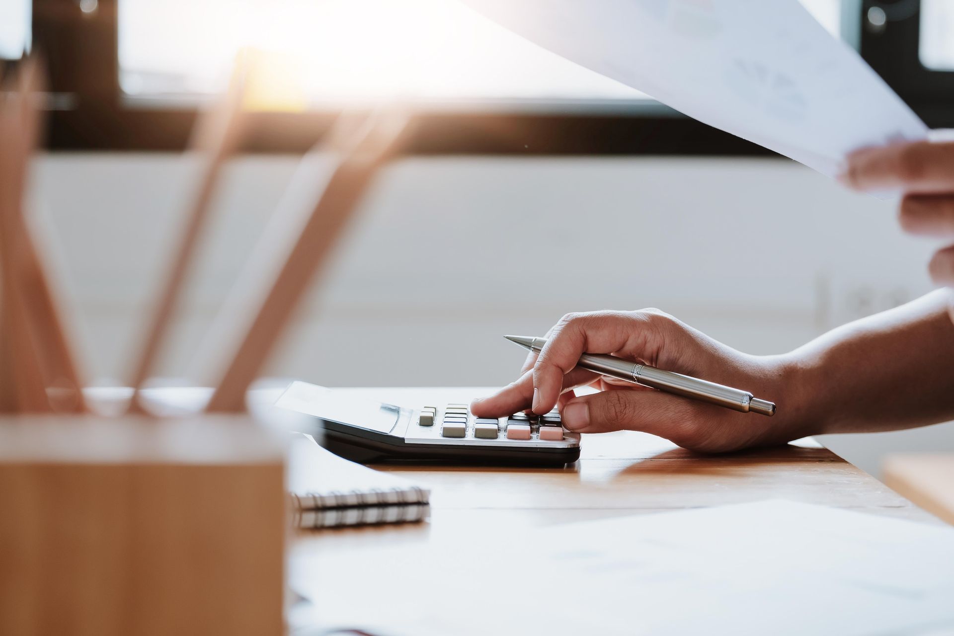 Person calculating with a calculator while holding papers at a desk, sunlit room.