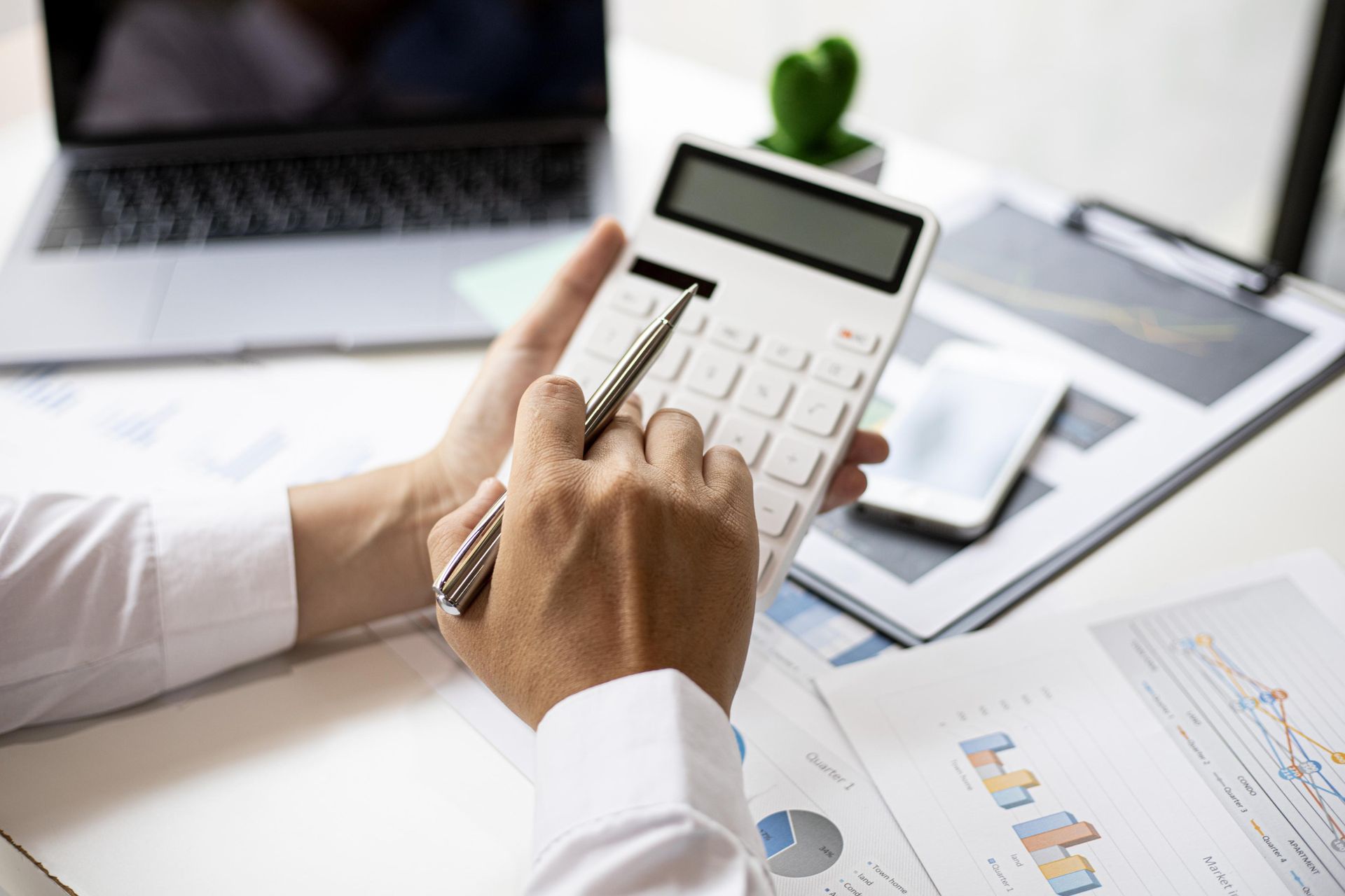 Person using a calculator with a pen, laptop, phone, and charts on a desk.