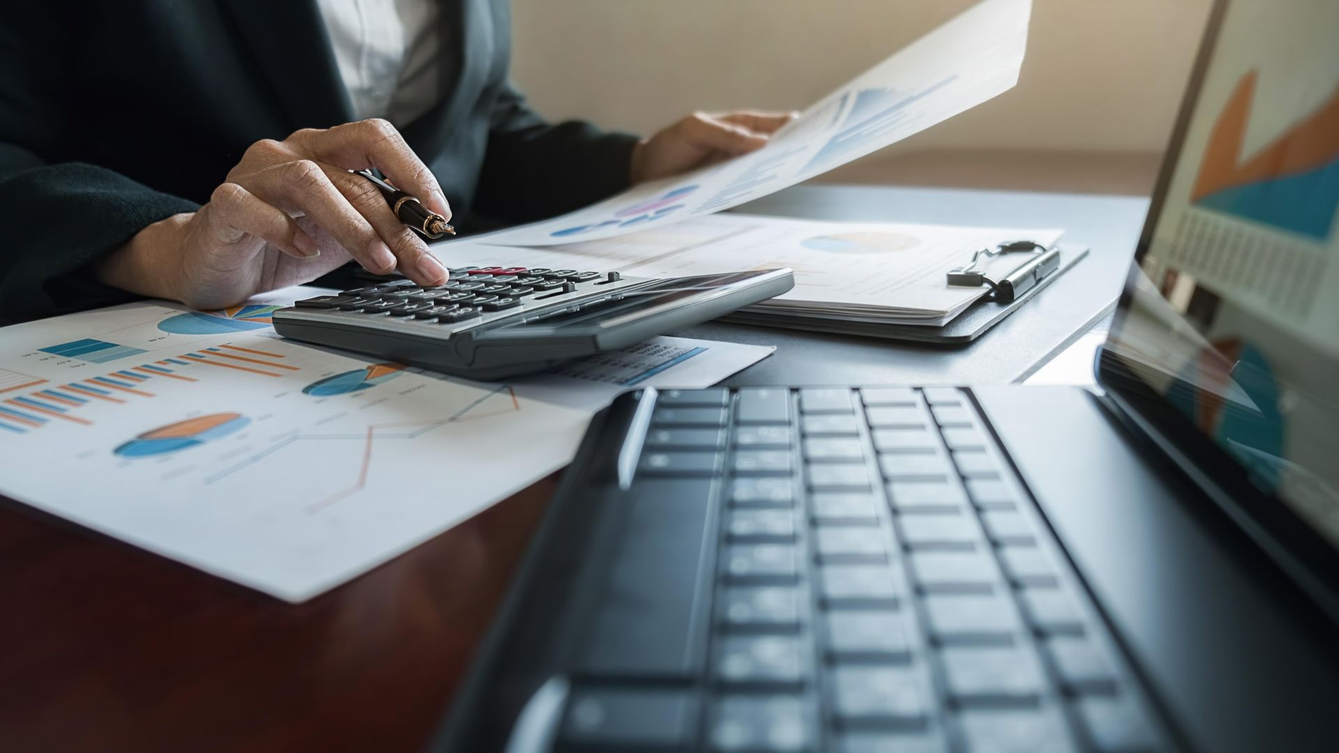 Person calculating with a calculator, looking at charts and laptop on a desk.