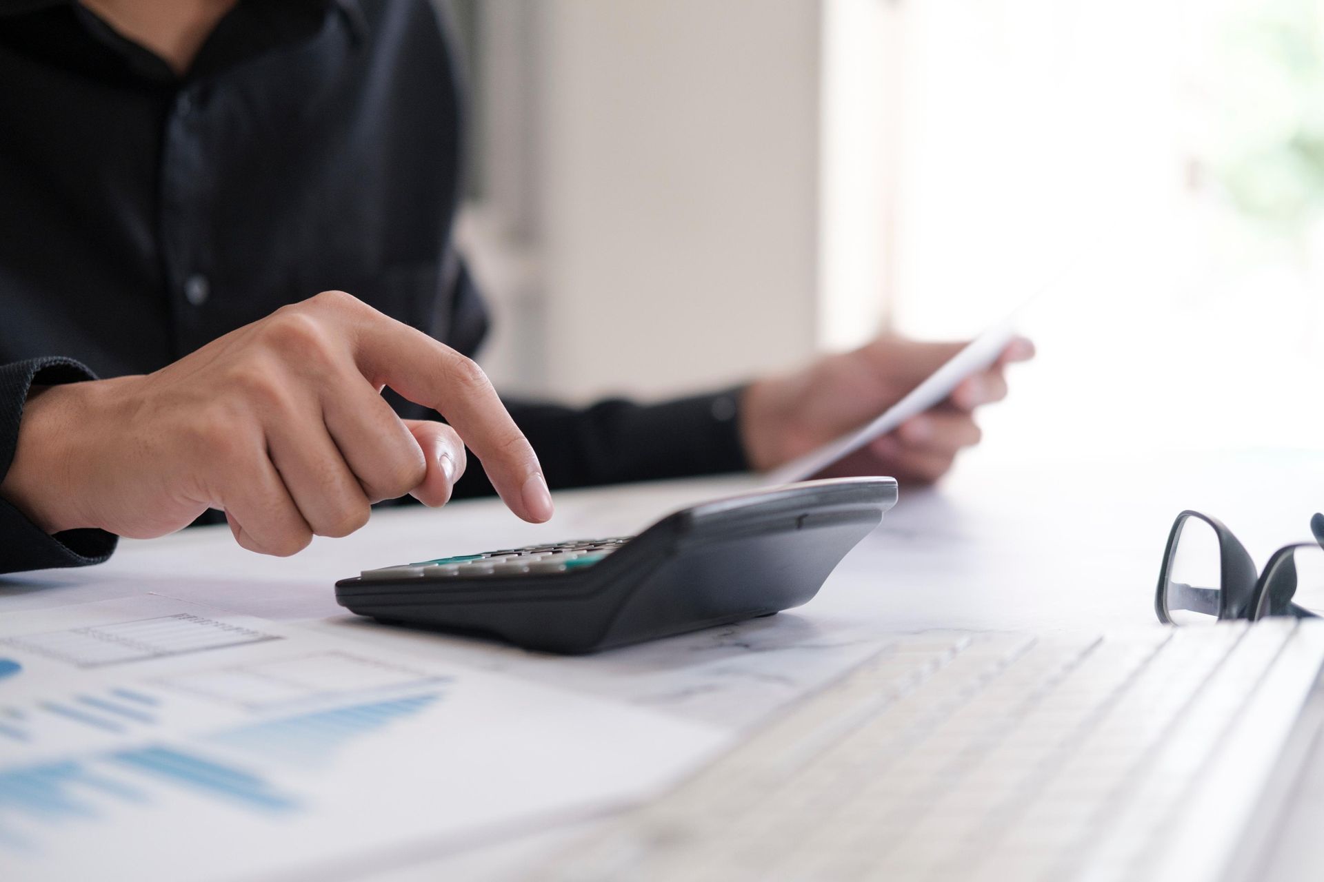 Person in black shirt using calculator, reviewing papers, with glasses on table and charts.