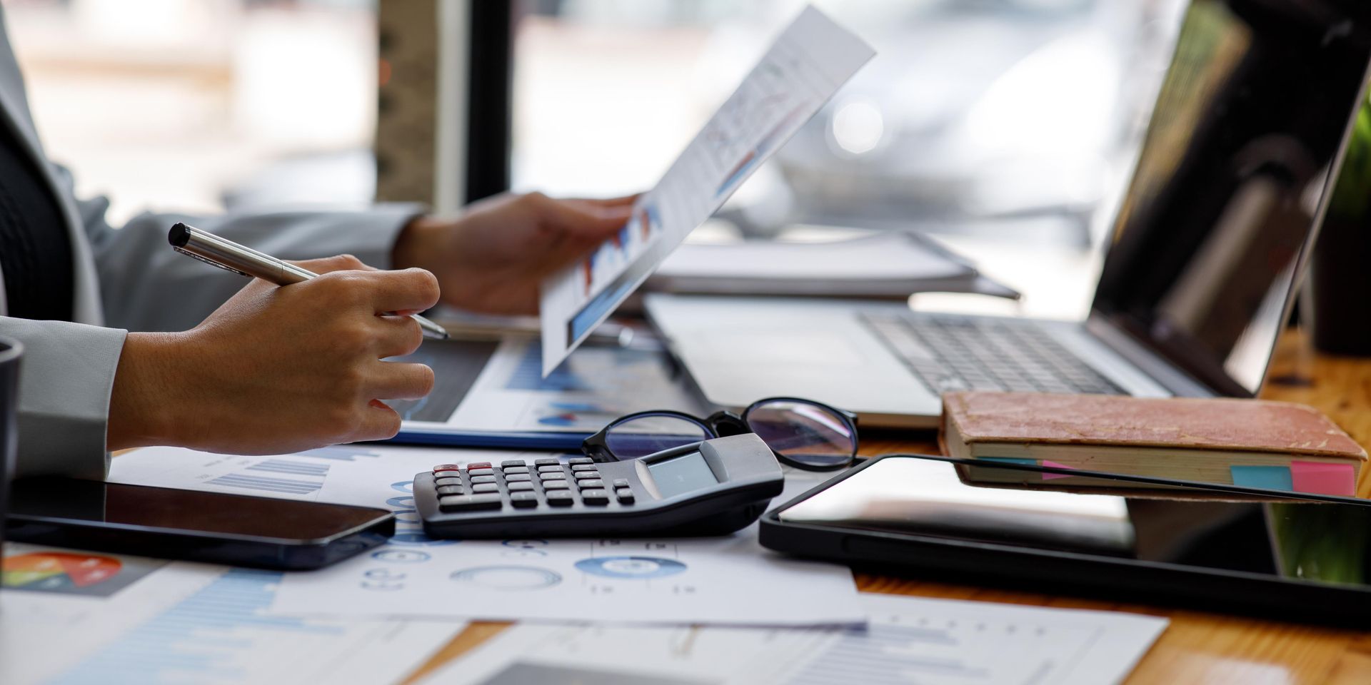 Person analyzing financial documents with a pen, calculator, laptop, and phone on a desk.