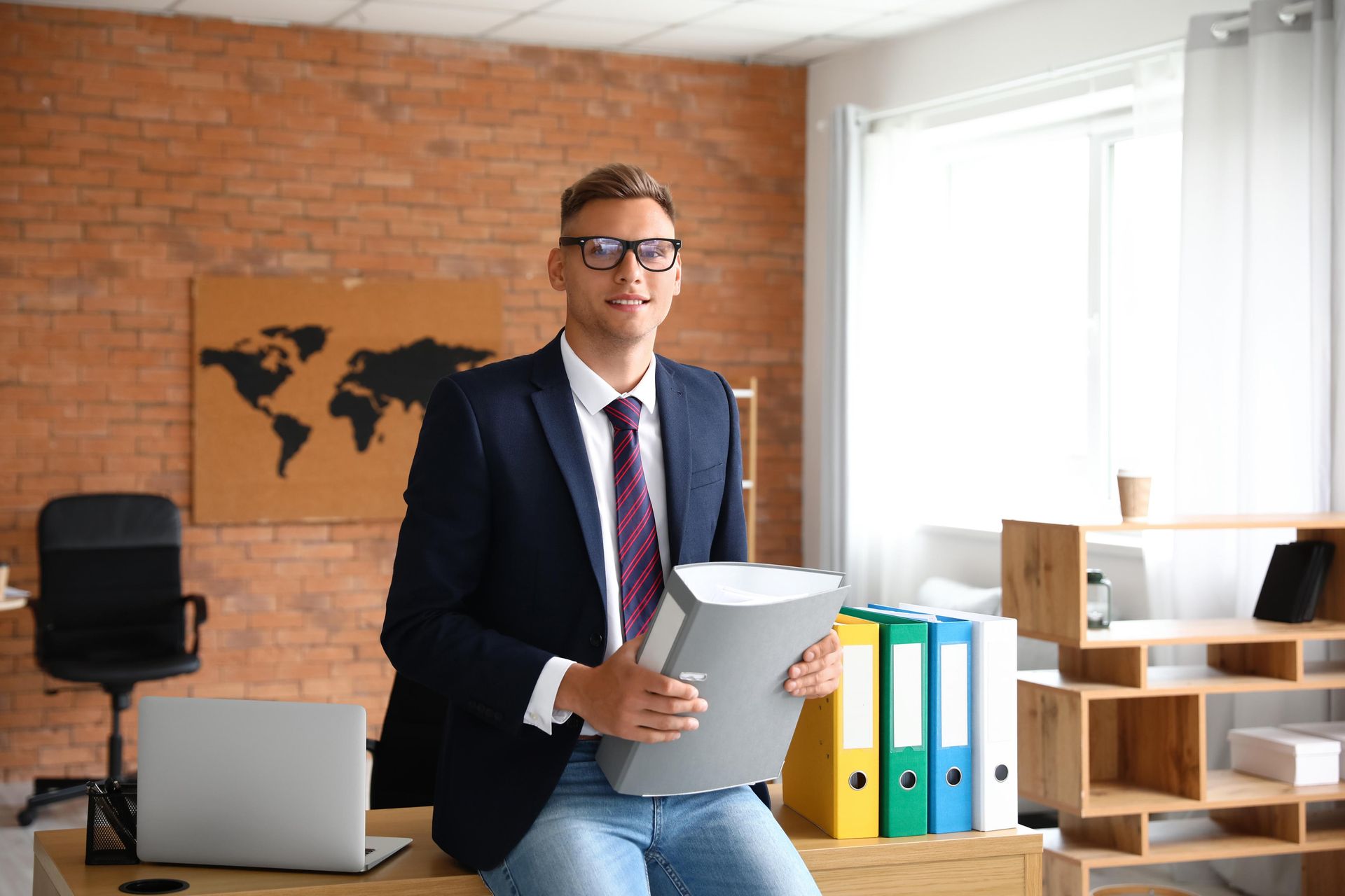 Man in a blazer and glasses sits on a desk holding a binder in a brick-walled office.