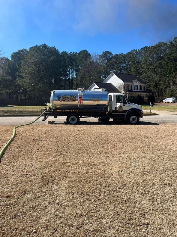 A tanker truck is parked in a gravel lot next to a house.