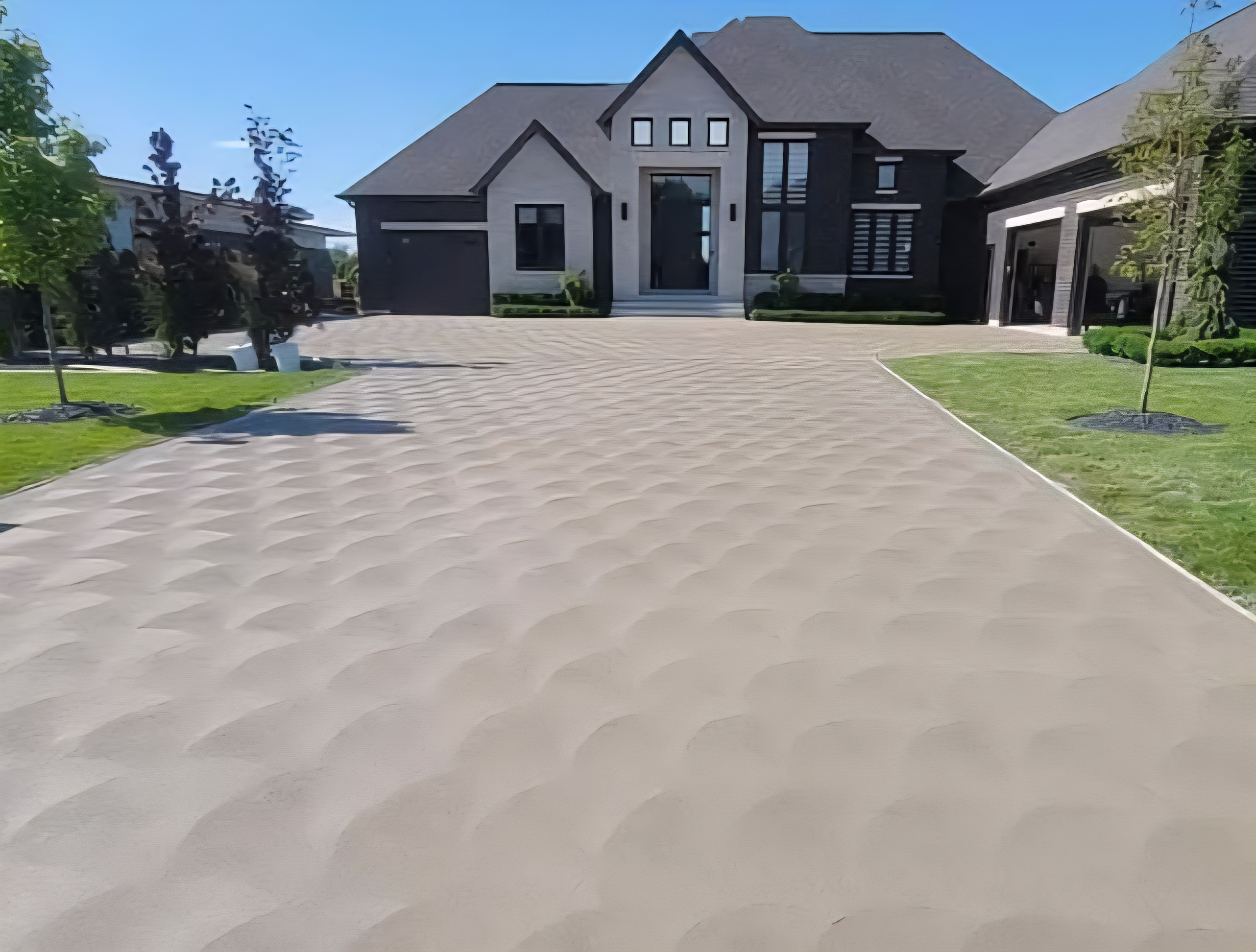 Beige textured driveway leading to a modern, dark-colored house with a manicured lawn.