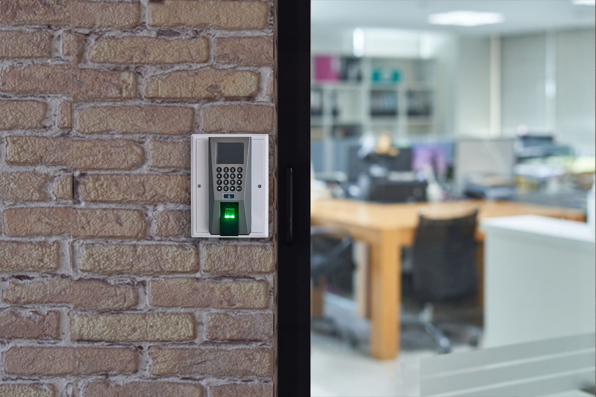 Wall-mounted keypad with fingerprint scanner beside a glass door leading into an office.