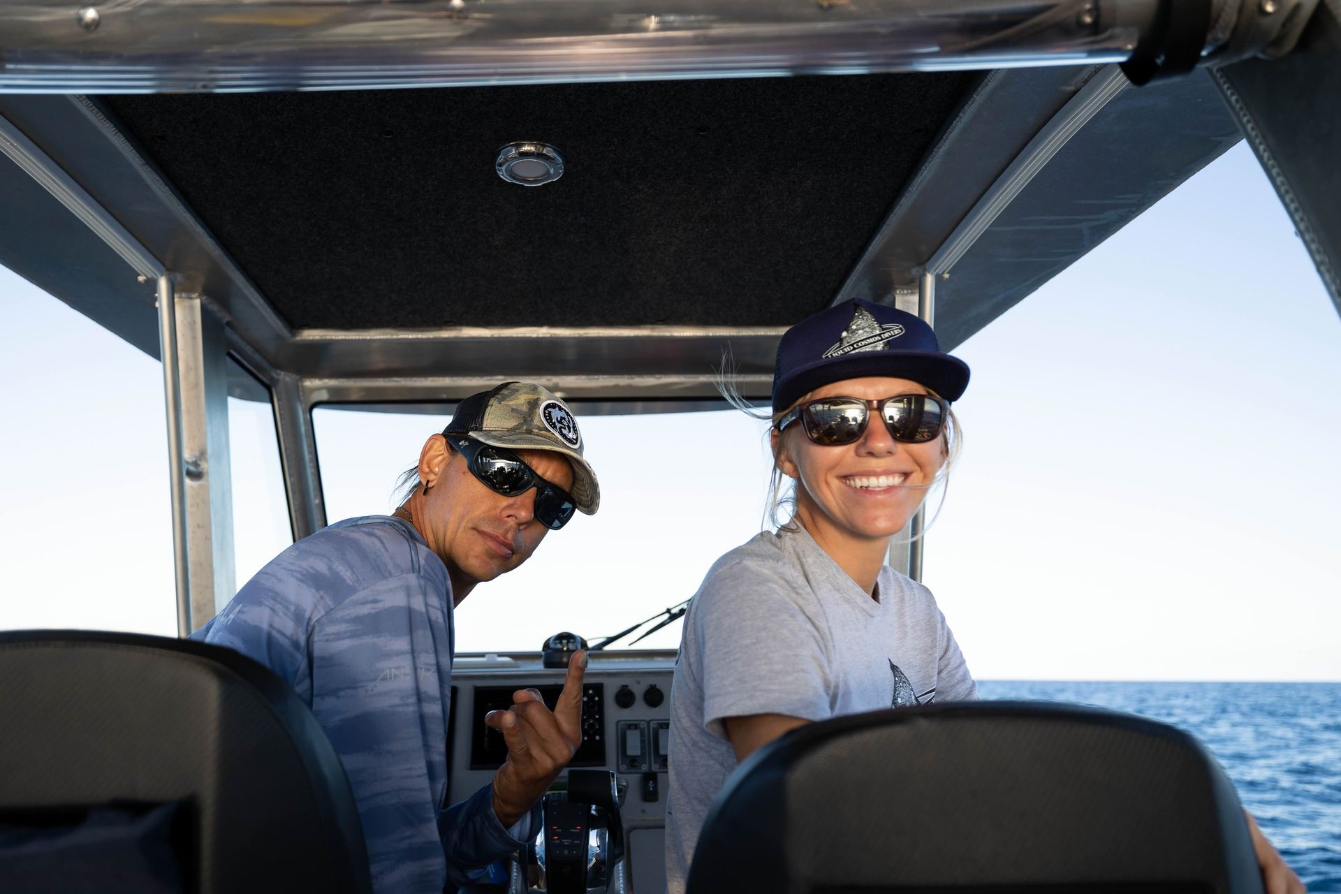 Two people wearing hats and sunglasses sit on a boat, smiling at the camera on open water.