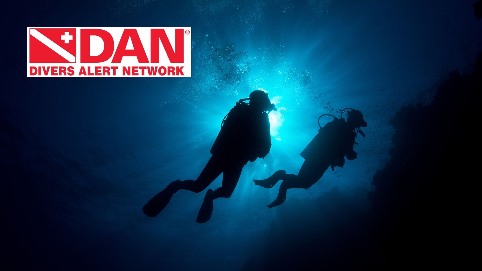 Two scuba divers silhouetted in blue underwater light beside a DAN Divers Alert Network logo
