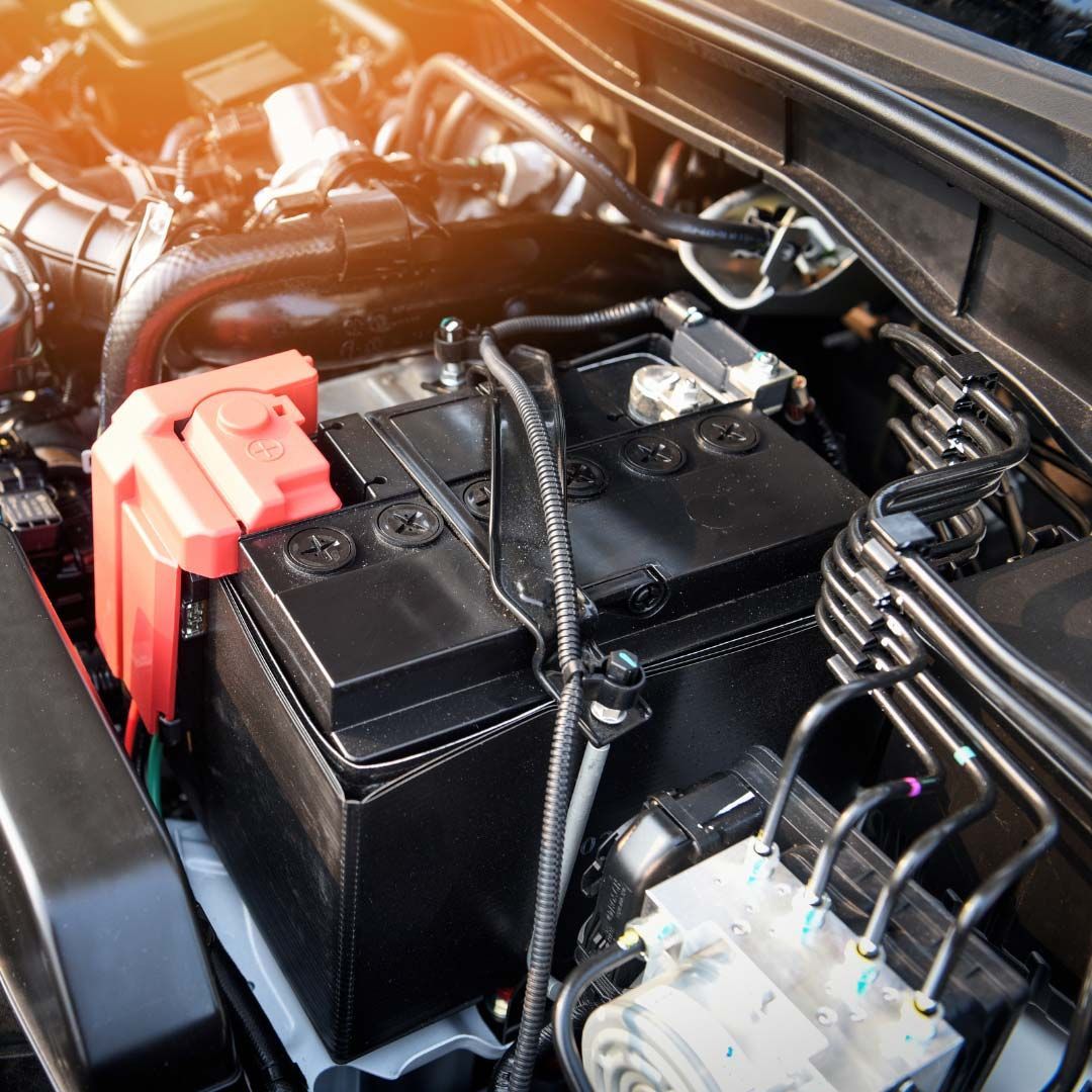 Car engine bay with a black battery, red terminal cover, and visible hoses and wiring
