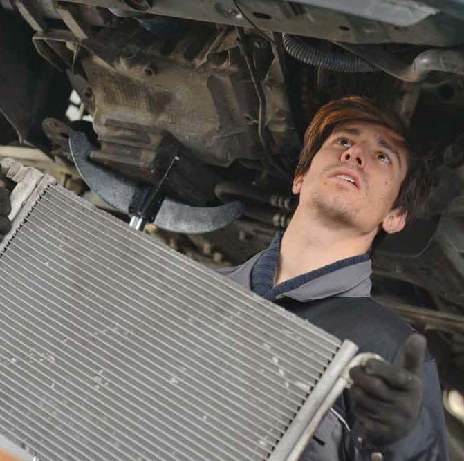 Mechanic inspecting a car’s radiator under the hood, wearing gloves and looking upward.