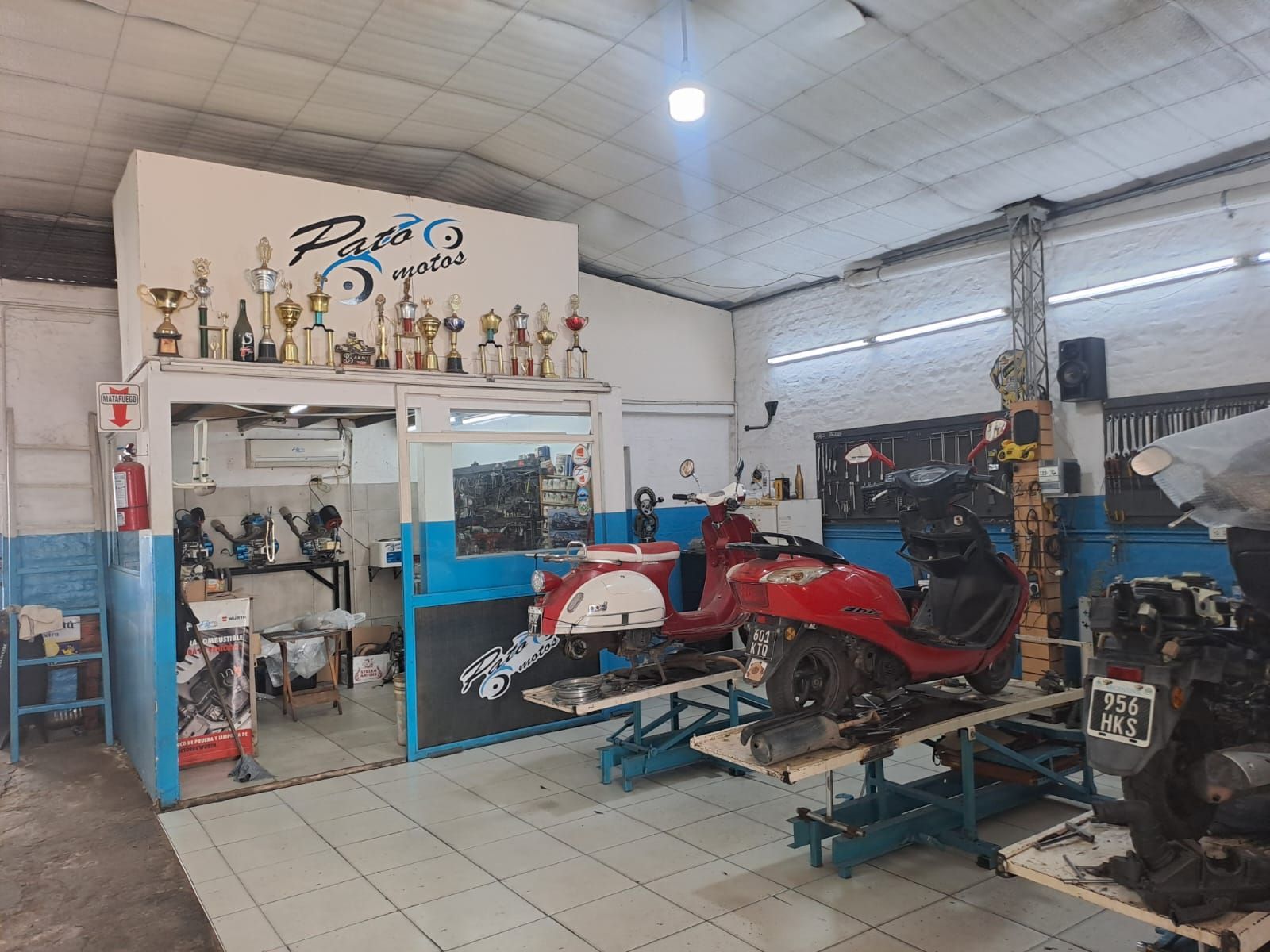 Taller mecánico con una motocicleta roja en un elevador, herramientas, trofeos y piezas en un garaje desordenado