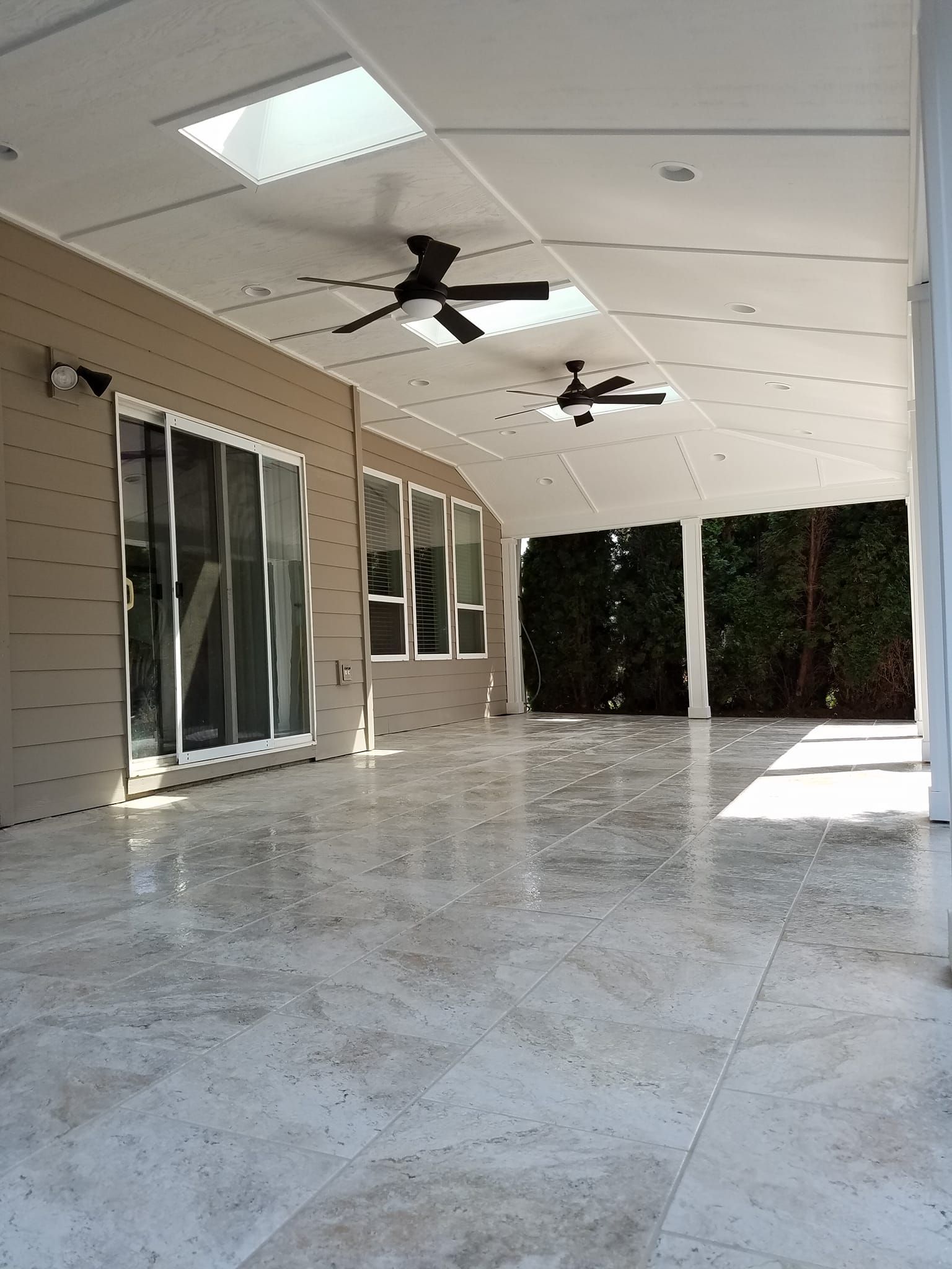 Covered patio with light-colored tile floor, tan siding, large windows, two ceiling fans, and a skylight.