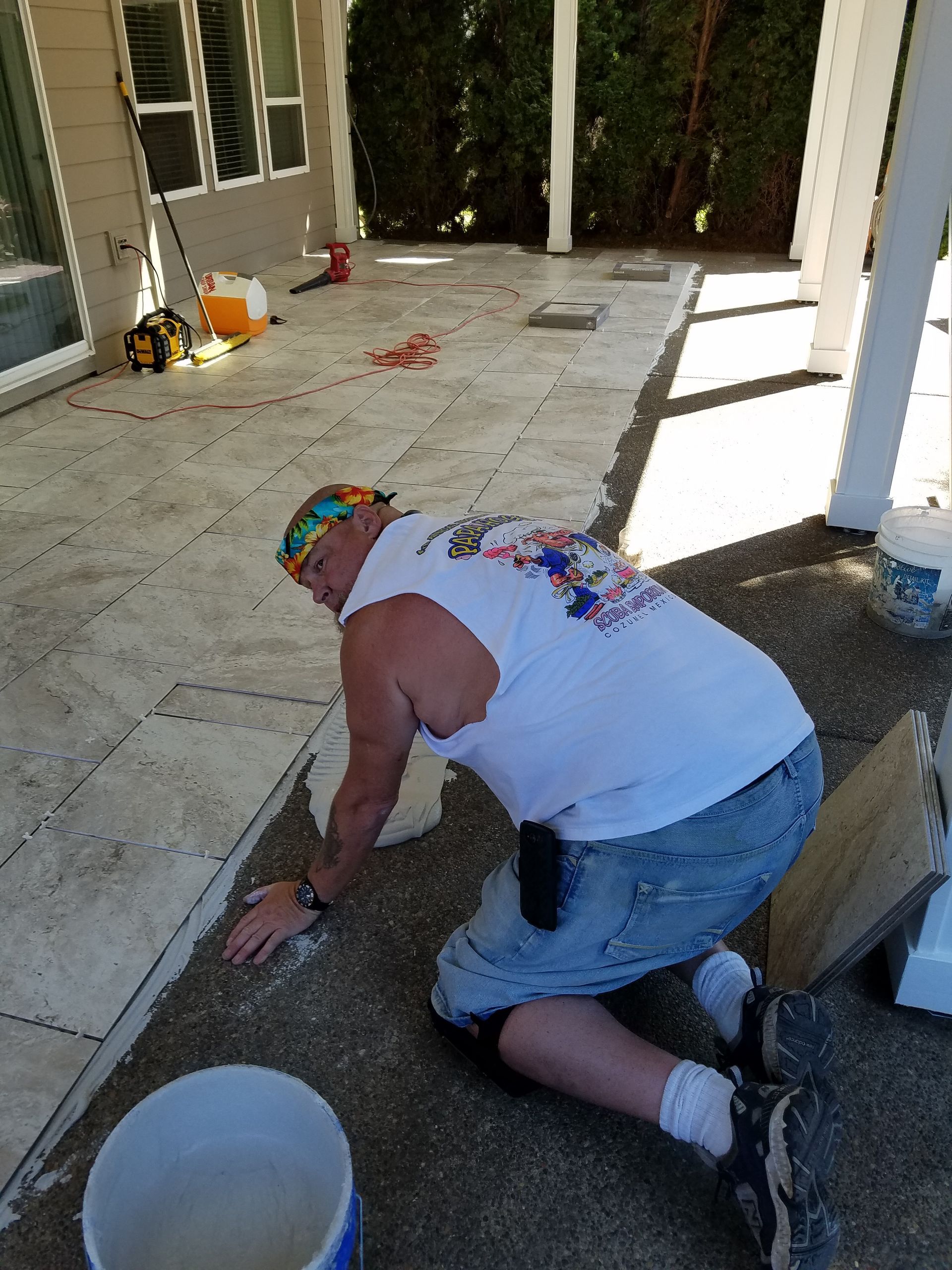 Man kneeling, working on patio; gray concrete, white pillars, sunny.