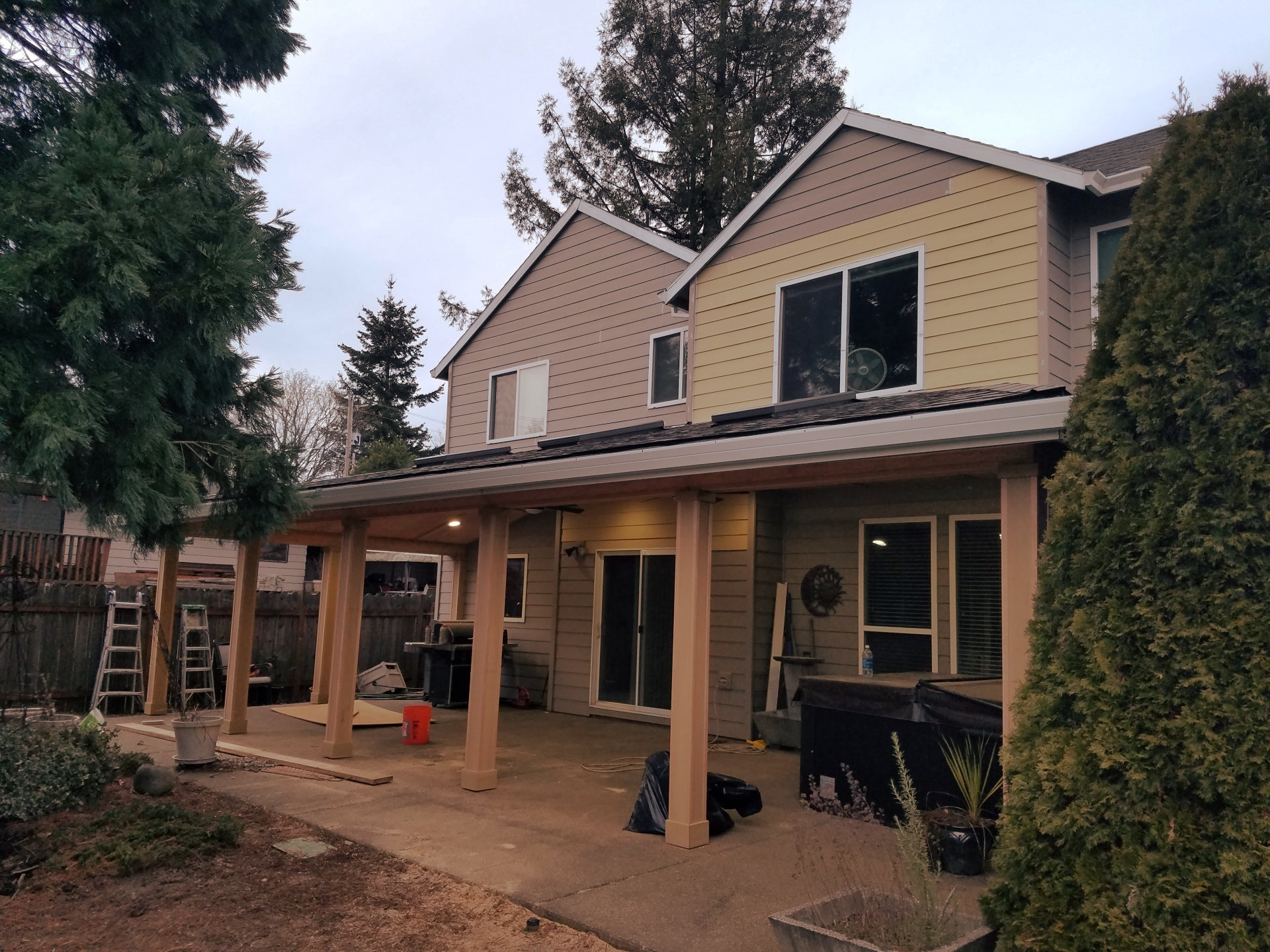 Backyard patio under construction with wooden beams and a partial roof, next to a two-story house.