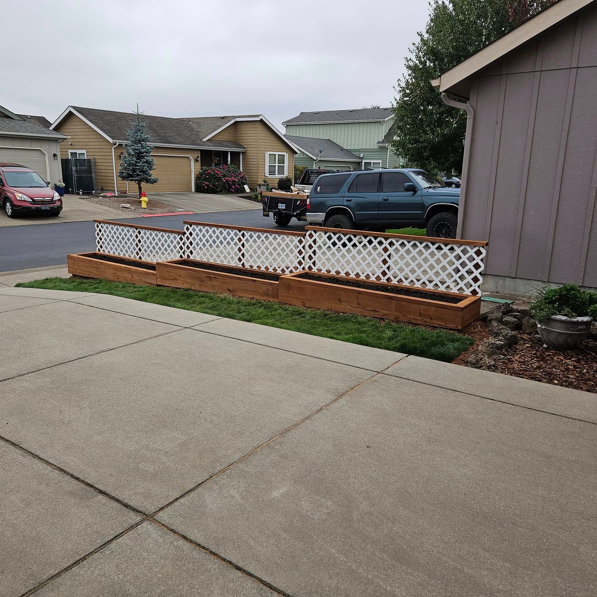 Front yard with a raised white lattice planter bed beside a concrete driveway in a suburban neighborhood.