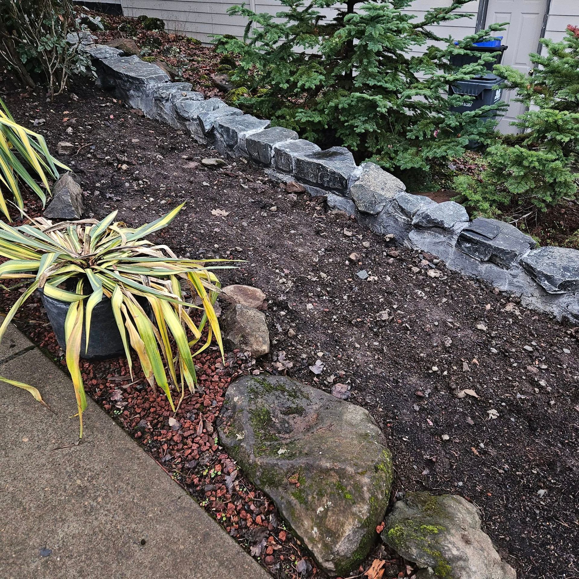 Rock garden with stepping stones, dark soil, variegated plants, and gray retaining blocks beside a walkway