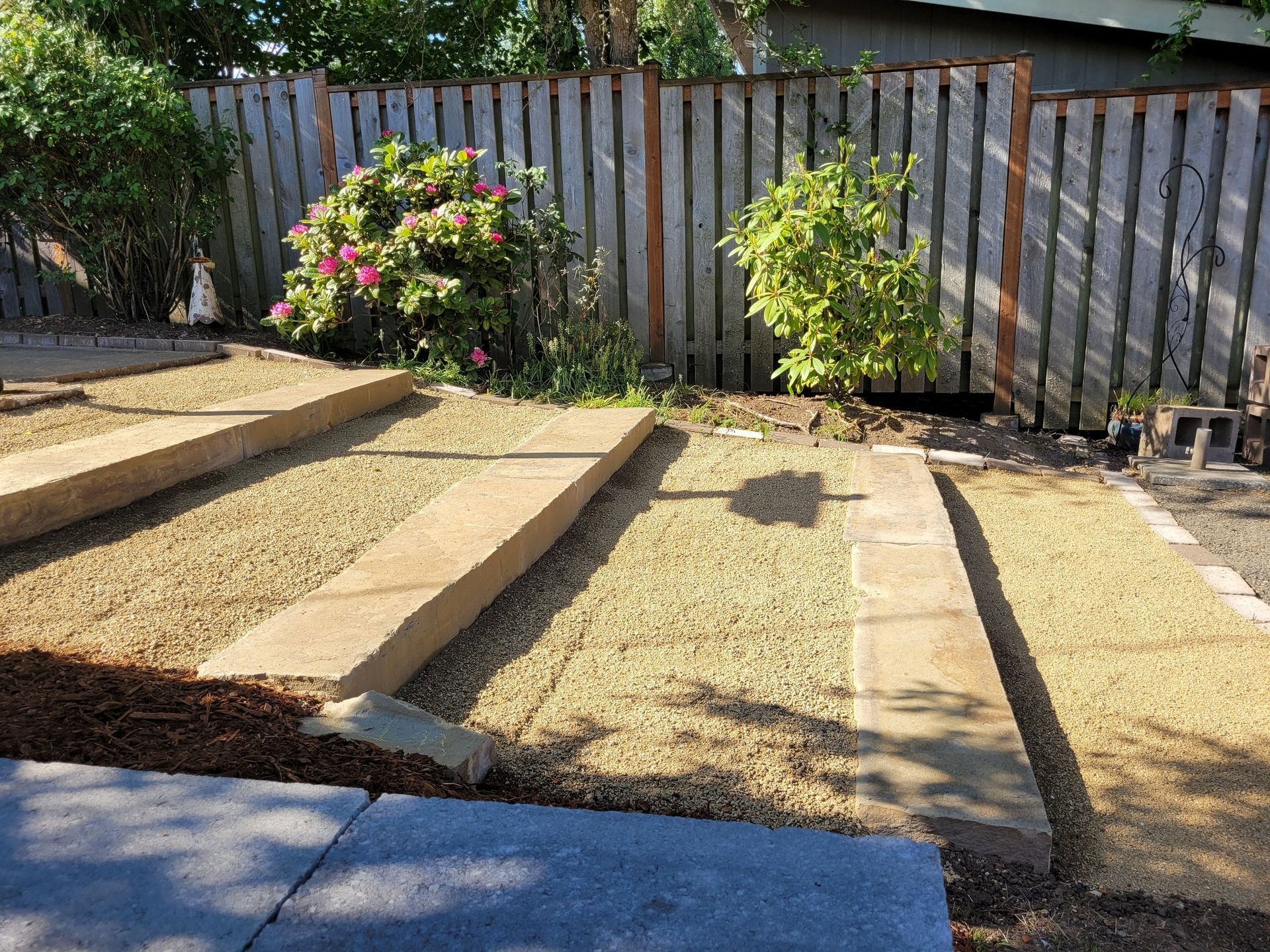 Raised garden beds in a sunny backyard with dry grass and flowering shrubs along a wooden fence