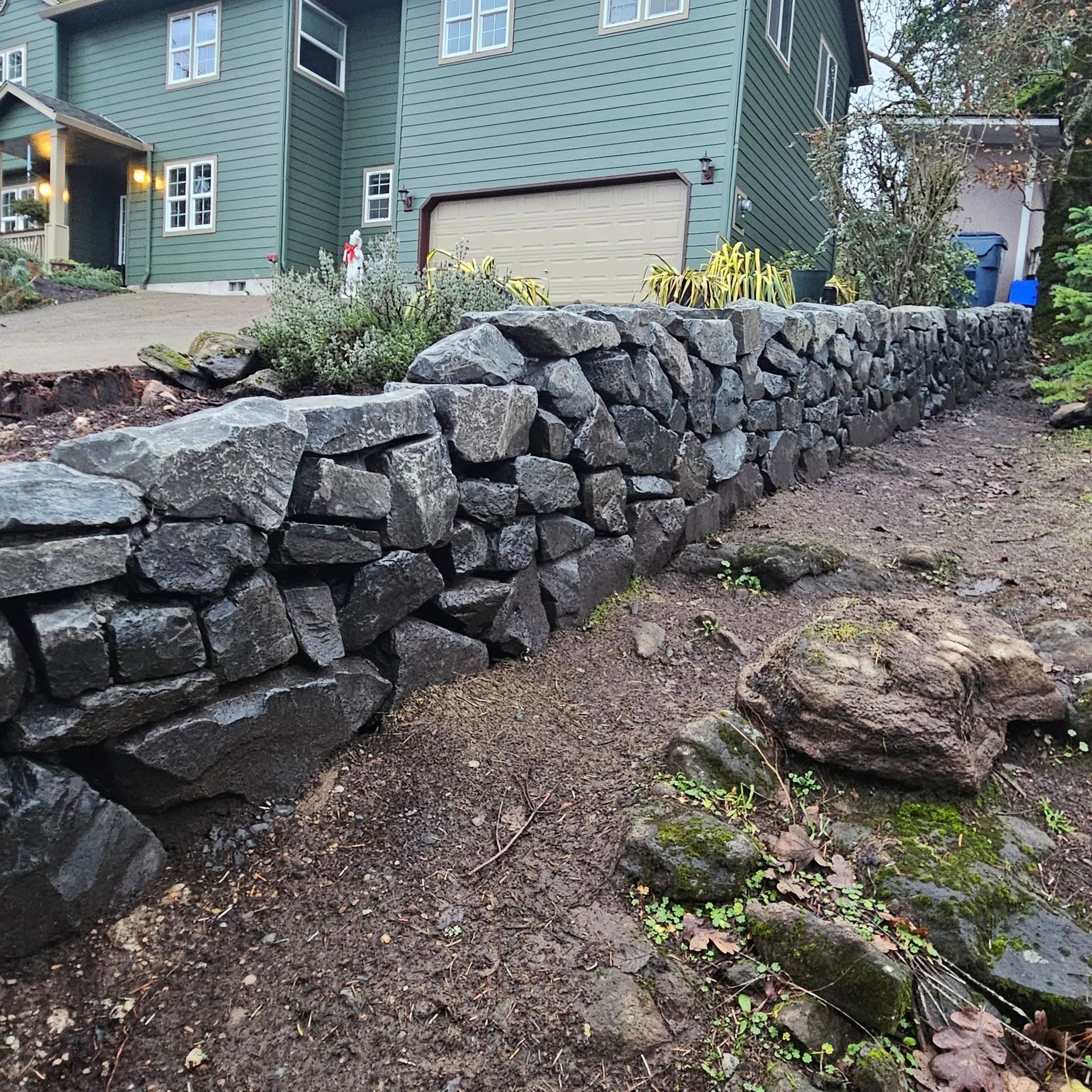 Stone retaining wall beside a green house, bordering a muddy sloped yard with rocks and plants.