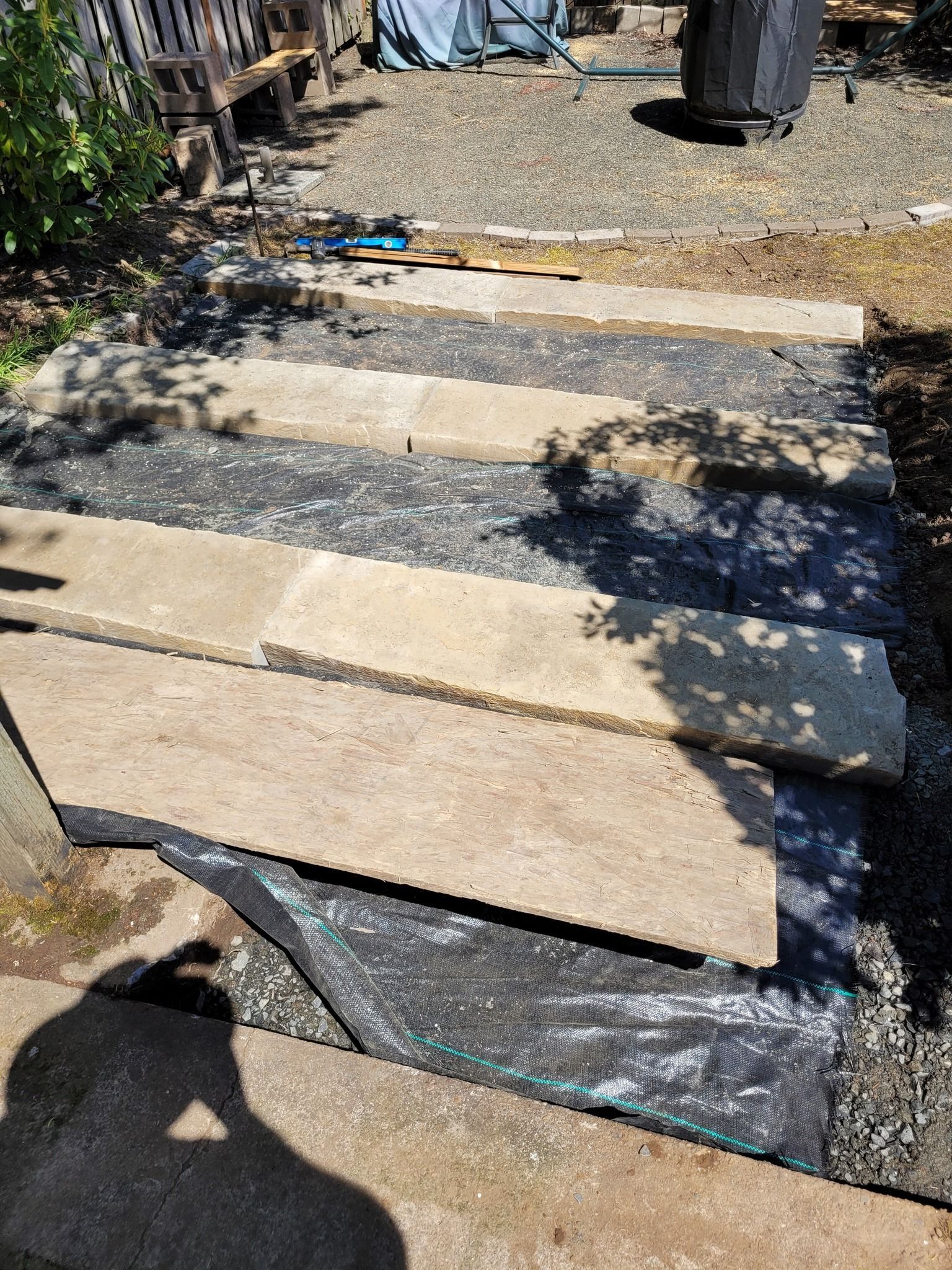 Stone pavers laid over gravel in a backyard walkway, with shadows and garden tools nearby