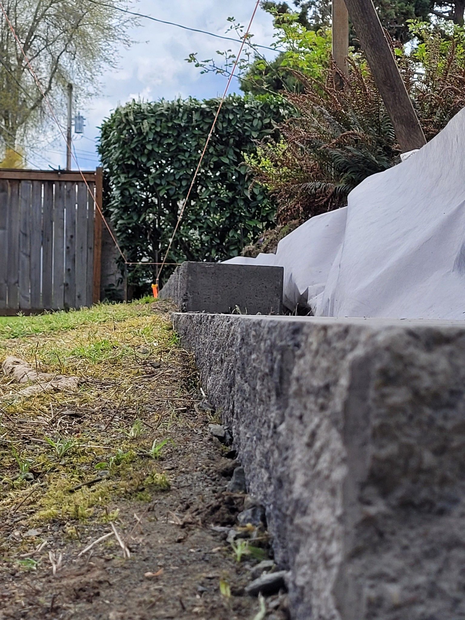 Backyard stone retaining wall beside a wooden fence, grass, and white tarp in daylight