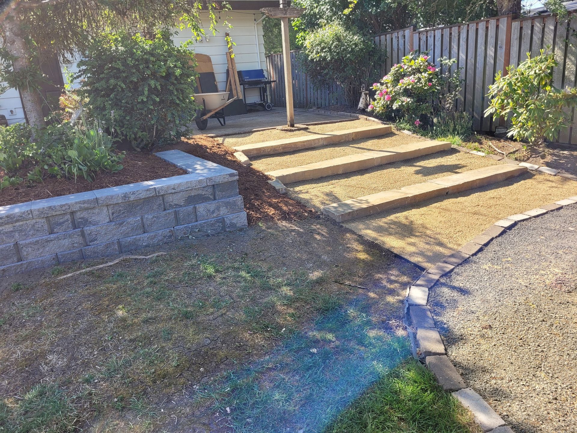 Outdoor steps beside a stone retaining wall and garden, leading to a shaded patio in bright sunlight